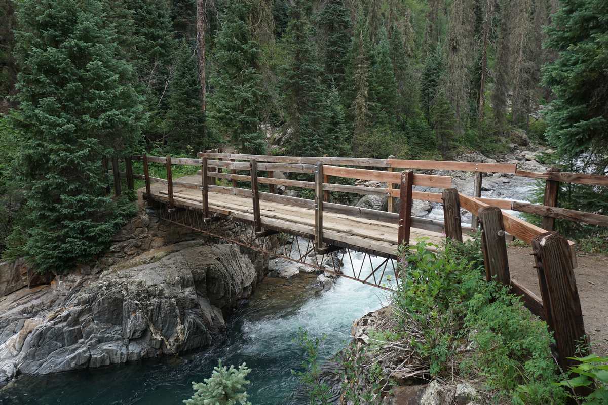 Wooden footbridge with log railings crossing Vallecito Creek between boulders, surrounded by evergreen forest, photographed before the October 2025 flood.