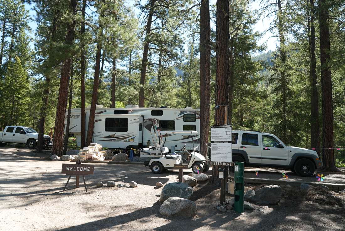 A camping trailer, camp-host golf cart, and a Register Here self-registration fee post at Vallecito Campground among tall pines