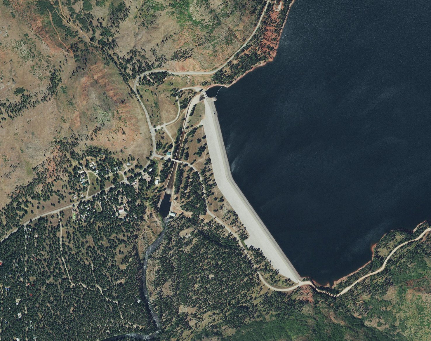 Vallecito Dam from directly overhead: the pale earthfill embankment, the concrete spillway chute beside its northern end running down to the Los Pinos River, and a band of bare shoreline around the reservoir