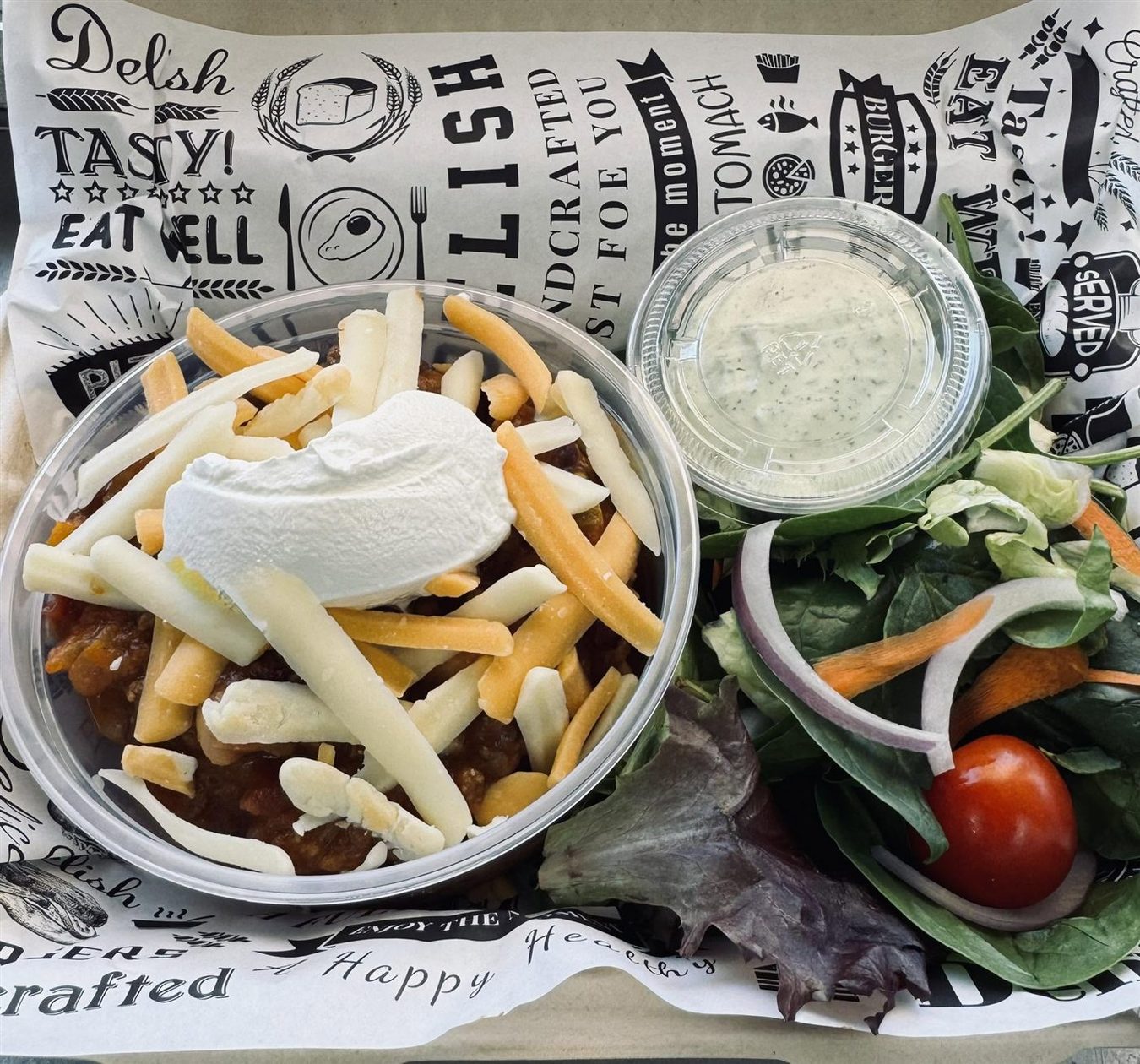 A bowl of chili with sour cream and crispy fries beside a side salad and ranch