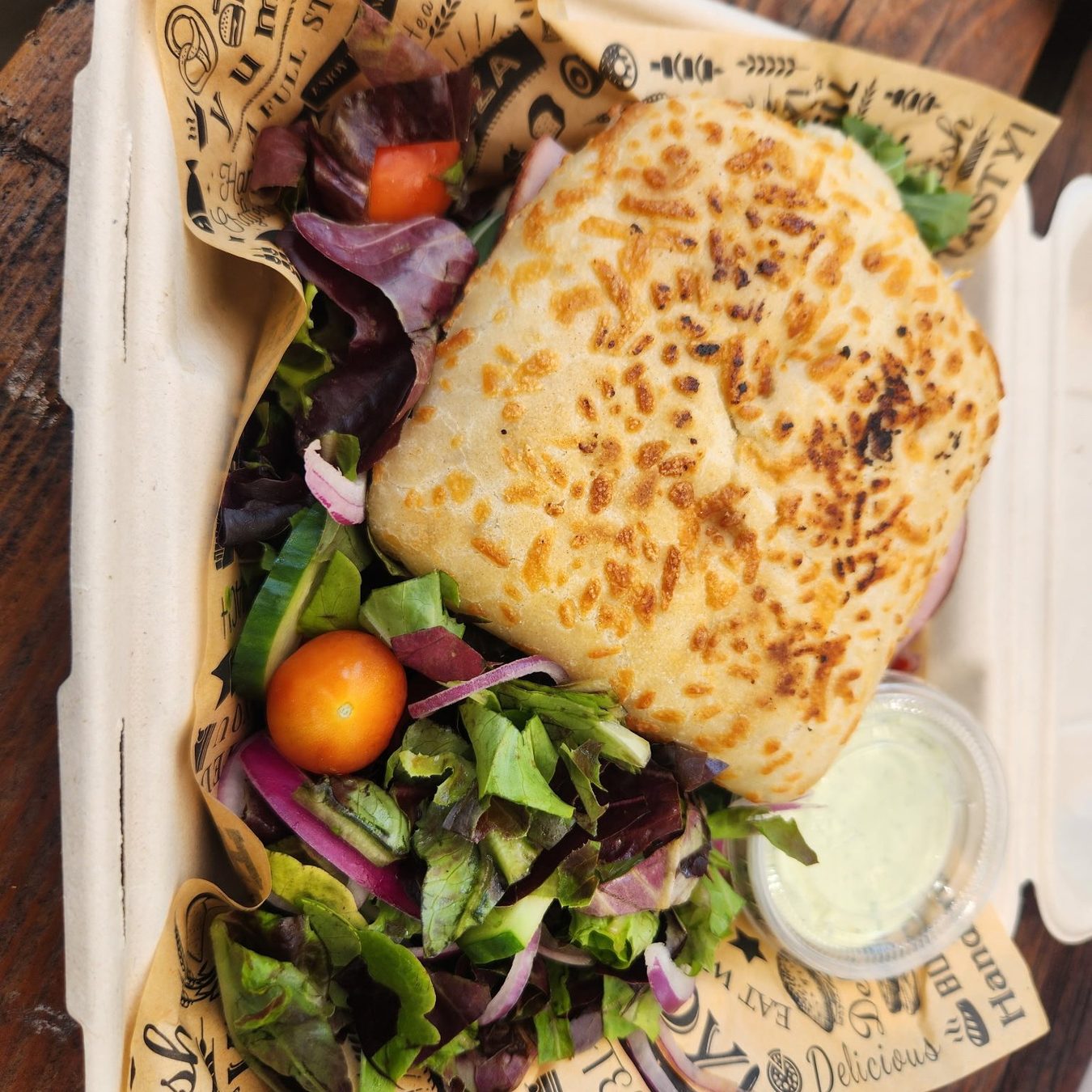 A toasted sesame-crusted sandwich in a takeout box with a green salad and dipping sauce