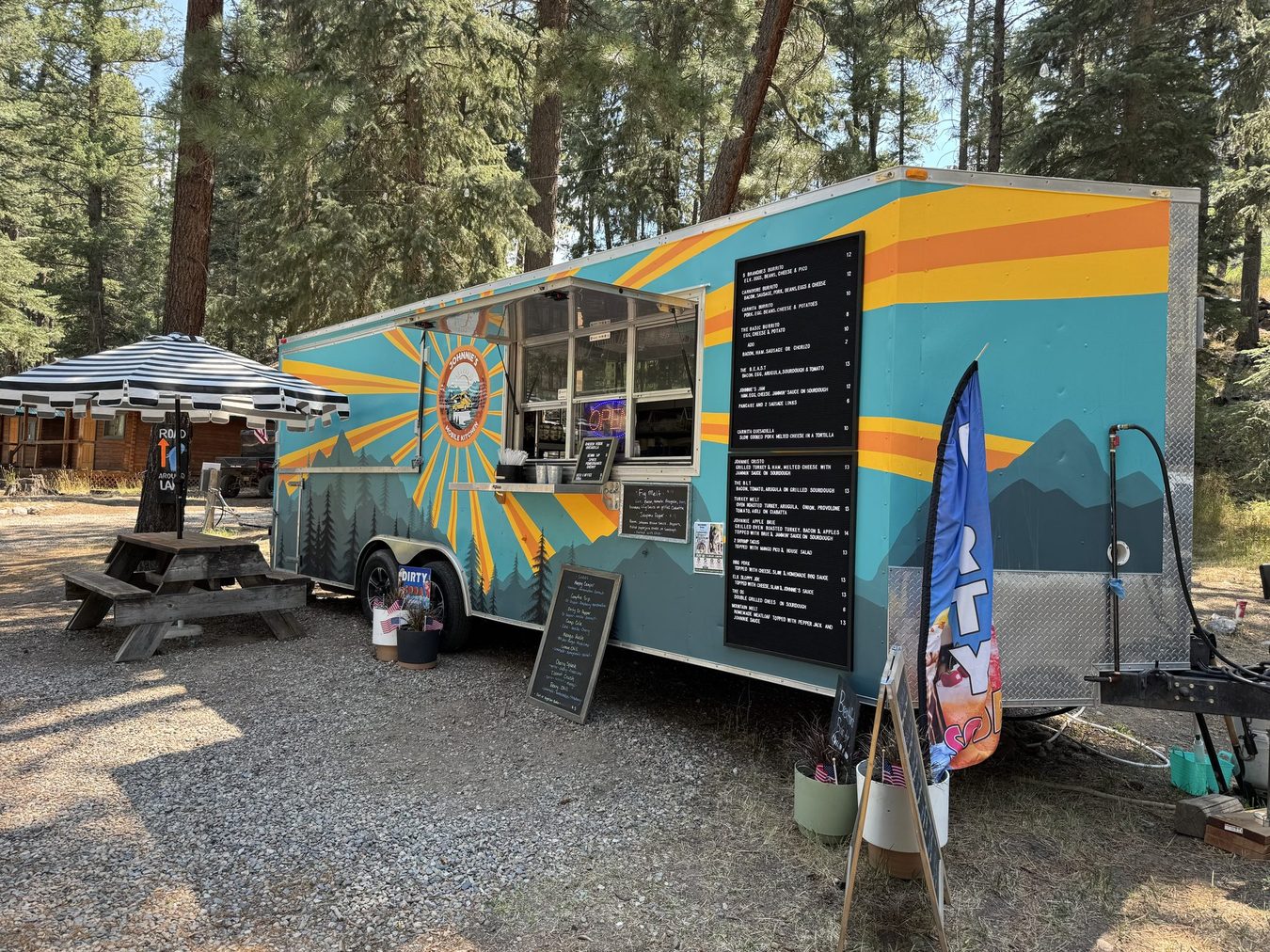 The teal Johnnie's trailer with its sunrise mural and menu boards parked under tall pines beside a picnic table